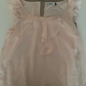Ljusrosa blus från Only - Söt ljusrosa blus från Only med volangdetaljer framtill. Blusen är ärmlös och har en rund halsringning. Perfekt för en somrig look.        Dock så är den klippt där nere men inget man tänker på