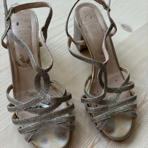 Glittriga sandaler med klack från Novita - Snygga glittriga sandaler från Novita med en bekväm blockklack. De har flera remmar som korsar foten och en justerbar rem runt ankeln. Perfekta för att lägga till lite glamour till din outfit. Något slitna, därav det låga priset. Nypris 700kr.