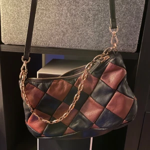 Rutig axelväska med kedjedetalj - Snygg axelväska med rutigt mönster i svart och brunt. Väskan har en kedjedetalj och dragkedja upptill. Perfekt för att ge din outfit en unik touch.