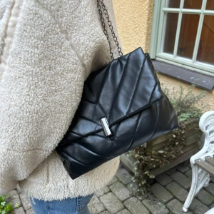 Svart axelväska - Snygg svart axelväska med kedjerem och stilrent spänne. Väskan har en modern design och är lätt att matcha med olika stilar.