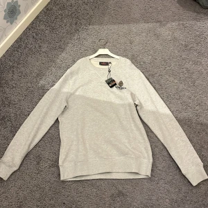 Grå sweatshirt från Morris - Säljer en stilren grå sweatshirt från Morris med rund hals och lång ärm. Tröjan har en broderad logga på bröstet och är perfekt för en avslappnad look. Helt ny med taggar kvar. Säljer för 900. Nypris 160. Pris kan diskuteras 