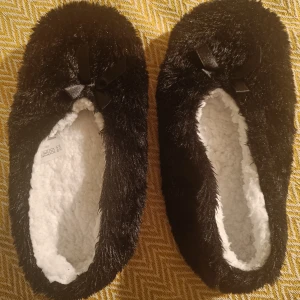 Svarta pälsiga tofflor med rosett - Mysiga svarta tofflor med pälsig utsida och mjuk, vit insida. De har en söt rosett på ovansidan och en grå sula. Perfekta för att hålla fötterna varma och bekväma hemma. Strl 39. Inget spec märke. 