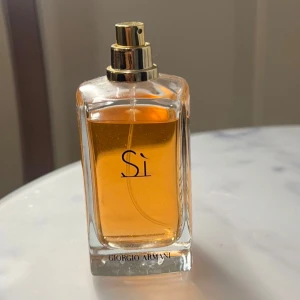 Sì Eau de Parfum från Giorgio Armani - Elegant parfymflaska med gyllene vätska från Giorgio Armani. Sì är en sofistikerad doft med inslag av vanilj och blommiga toner. Flaskan har en stilren design med en svart kork och guldiga detaljer. Locket saknas.
