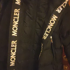 Svart dunjacka från Moncler - Snygg svart dunjacka från Moncler med logotypband längs dragkedjan. Jackan är designad för lätt sport och har vattenavvisande egenskaper samt bra andningsförmåga. Den har en vattentät dragkedja och vindtäta detaljer. Dock en reva som behöver sys men är inte svårt och går att få fint se bild därav lågt pris 