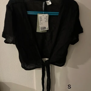 Svart knytbolero från Gina Tricot - Snygg svart bolerotopp från Gina Tricot med knytning framtill. Perfekt för att lägga till en extra touch till din outfit. Kortärmad och i ett lätt material.