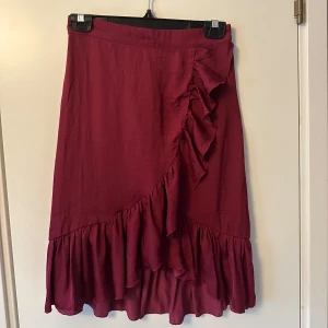 Vinröd kjol från NA-KD - Snygg vinröd kjol från NA-KD med volangdetaljer. Kjolen har en omlott-design och är perfekt för att ge en elegant touch till din outfit. Den är tillverkad i ett mjukt material som ger en skön passform.