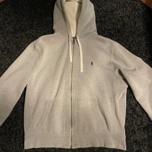 Ralph Lauren Zip Hoodie - Säljer en stilren grå zip hoodie från Polo Ralph Lauren med en röd logga på bröstet🔥 Perfekt nu till vintern❄️ Tröjan är i väldigt fint skick förutom lite slitningar i luvan som nu är lagade! Storlek XL men passar även L. Hör av dig vid funderingar/ Karl!
