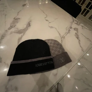 Två mössor från Louis Vuitton (Äkta) - Säljer två stilrena mössor från Louis Vuitton. Den ena är svart med grå rand och broderad logotyp, den andra är grå med ett diskret mönster och logotyp. Perfekta för att hålla värmen med stil.