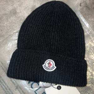 Svart ribbad mössa från Moncler - Snygg svart ribbad mössa från Moncler med broderad logotyp framtill. Perfekt för kyliga dagar och ger en stilren look.