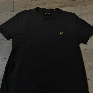 Svart t-shirt från Lyle & Scott - Snygg svart t-shirt från Lyle & Scott med deras ikoniska logga broderad på bröstet. Perfekt för en stilren och avslappnad look. Tillverkad i mjukt material för extra komfort.