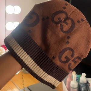 Brun mössa med GG-mönster - Snygg brun mössa från Gucci med det ikoniska GG-mönstret. Mössan har ribbad kant i mörkbrunt och beige, vilket ger en stilren kontrast. Perfekt för att hålla värmen med stil. Köpt för 4000kr här på Plick som oanvänd används inte längre sparsamt använd 