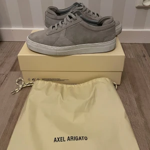 Grå sneakers från Axel Arigato - Snygga grå sneakers från Axel Arigato med stilren design. Skorna har en tjock vit sula och är tillverkade i mocka. Skorna är endast använd 1 gång! Påse, samt sko kartongen ingår, kontakta för bättre bilder 