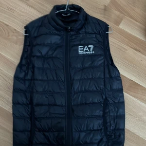 Svart quiltad dunväst från Emporio Armani - Säljer en snygg svart quiltad dunväst från Emporio Armani EA7. Västen har en dragkedja framtill och är perfekt för både höst och vår. Den är i nyskick och har en stilren design med logotypen tryckt på bröstet. Pris ej hugget i sten