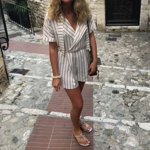 Maje Jumpsuit Small - En randig jumpsuit med V-ringning och korta ärmar. Den har en knäppning framtill och en dragsko i midjan. Jumpsuiten är i en vit basfärg med röda och svarta ränder.  Bra skick, några få trådar som gått upp se bild