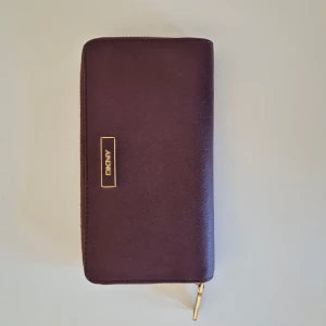Plånbok DKNY - Plånbok från märket DKNY. Använd några gånger. Vinröd färg. Flera fickor insidan för korthållare, mynt, sedlar eller mobil. Rymlig och fräsh!  19 cm lång och 10 cm hög.