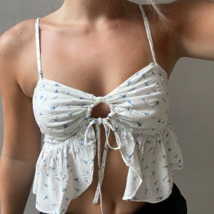 Vit blommig linnetopp med knytning - Säljer en superfin vit linnetopp med små blå blommor. Toppen har smala axelband och en söt knytning framtill som ger en liten öppning. Den är perfekt för skola och varmare dagar och har en luftig och bekväm passform.