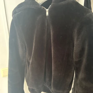 Svart teddyjacka med dragkedja - Mysig svart teddyjacka med dragkedja framtill. Perfekt för kyliga dagar när du vill vara både varm och stilren. Jackan har en huva och långa ärmar, vilket gör den extra bekväm och praktisk.