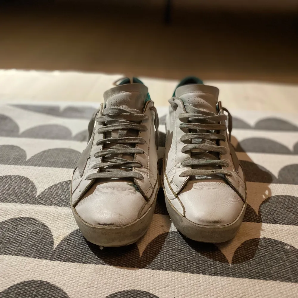 Säljer ett par vita golden goose med gröna detaljer. De har en klassisk design med snörning och en stjärna på sidan. Skorna är i skinn och har en lite sliten look som ger dem en cool vintage känsla. Perfekta för både vardag och fest!. Skor.