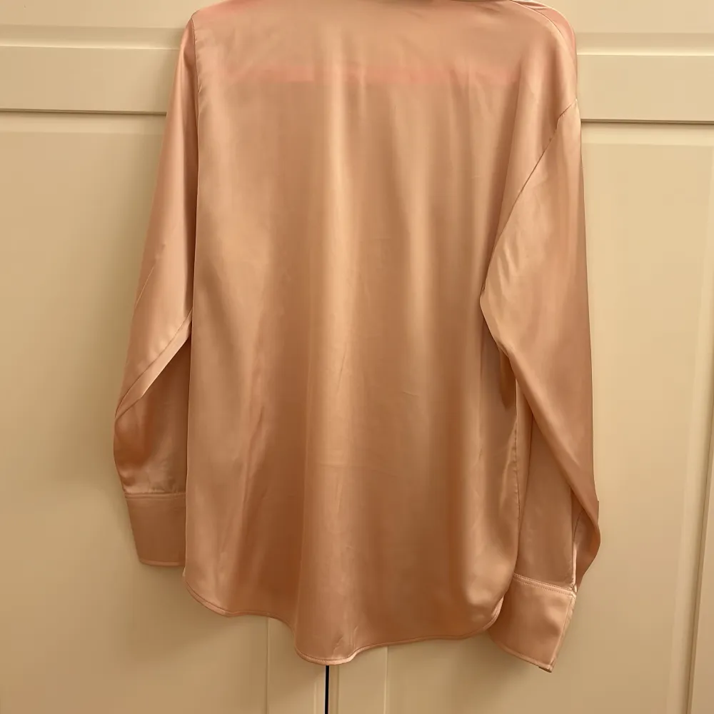 Säljer en elegant beige blus i satin från H&M. Den har en mjuk och glansig yta med långa ärmar och en klassisk krage. Blusen har en dold knäppning framtill som ger en stilren look. Perfekt för både vardag och fest!. Skjortor.