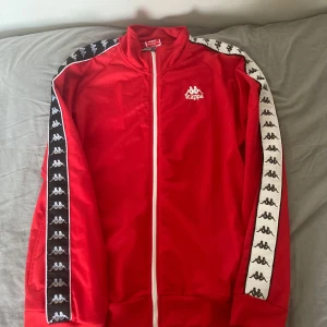 Röd träningsjacka från Kappa - Säljer en snygg röd träningsjacka från Kappa i storlek XXL. Jackan har en klassisk design med vita detaljer och Kappas logga längs ärmarna. Den är långärmad och har en dragkedja framtill. Perfekt för både träning och vardagsbruk!