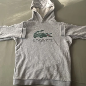 Grå Lacoste hoodie i storlek 164cm - Detta är en grå Lacost hoodie i storleken 164cm. Den har tryck på framsidan och även att de står lacost under trycket i svart färg. Denna hoddie har även inga snören på luvan. Den är skön och lätt.
