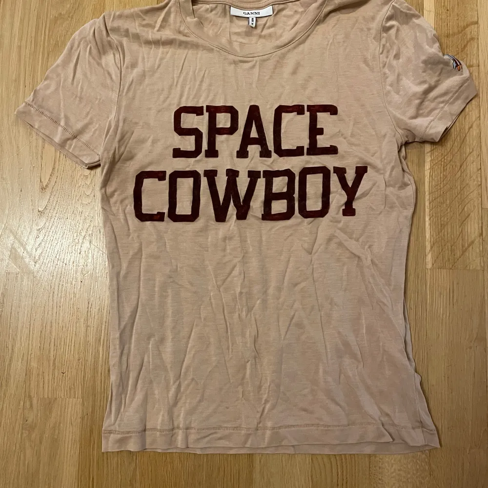 Säljer en supercool beige t-shirt från Ganni med texten 'Space Cowboy' i rödbrunt på framsidan. Den har korta ärmar och en liten broderad rymdraket på ena ärmen. Perfekt för en avslappnad och trendig look!. T-shirts.