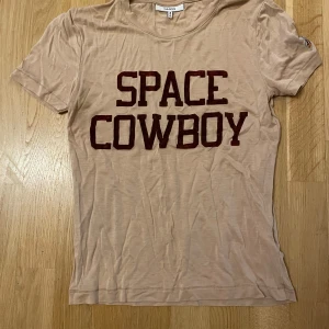 Beige t-shirt från Ganni med texten 'Space Cowboy' - Säljer en supercool beige t-shirt från Ganni med texten 'Space Cowboy' i rödbrunt på framsidan. Den har korta ärmar och en liten broderad rymdraket på ena ärmen. Perfekt för en avslappnad och trendig look!