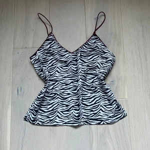 Zebramönstrad linnetopp från Bik Bok - Säljer en supercool zebramönstrad linnetopp från Bik Bok i storlek XS. Toppen har tunna axelband och en snygg v-ringning. Den är gjord av 100% polyester och har en kontrasterande vinröd kant som ger en extra touch. Perfekt för utekväll!