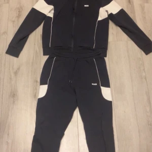 Svart träningsset från BOSS - Säljer ett stilrent svart träningsset från BOSS med vita detaljer. Setet består av en långärmad tröja med dragkedja och matchande byxor. Perfekt för en sportig look eller avslappnad dag. Köpt för 4000 kr. Ny skick, använda få gånger. 