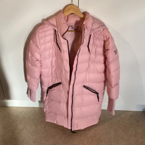 Rosa dunjacka från Reebok - Säljer en snygg rosa dunjacka från Reebok med huva och dragkedja. Jackan har långa ärmar och praktiska fickor med dragkedjor. Perfekt för kyliga dagar.