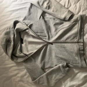 Grå zip hoodie från Sail Racing - Säljer en stilren grå hoodie från Sail Racing med dragkedja framtill och en liten ficka med dragkedja på bröstet. Perfekt för en avslappnad look. Huvan ger extra komfort och värme.