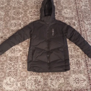 Svart dunjacka från Sail Racing - Säljer en svart dunjacka från Sail Racing med huva och dragkedja. Jackan är stilren, passar med vilka kläder som helst, och är perfekt för kyliga dagar.