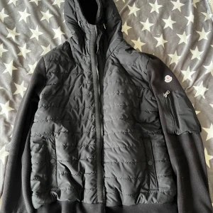 Svart hybrid jacka från Moncler - Snygg svart jacka från Moncler med quiltad design och dragkedja. Jackan har en hög krage och praktiska fickor med dragkedja på ärmen. Perfekt för kyligare dagar. Från liknande sida som panda sidan