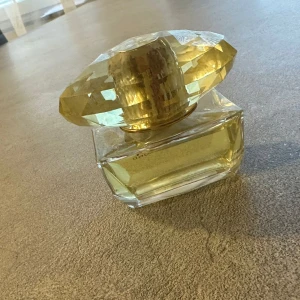 Guldfärgad parfymflaska - Elegant parfymflaska från vercace  med en guldfärgad, facetterad kork som liknar en diamant. Flaskan är genomskinlig och innehåller en ljusgul vätska. Perfekt för den som vill ha en lyxig touch på sitt parfymbord.