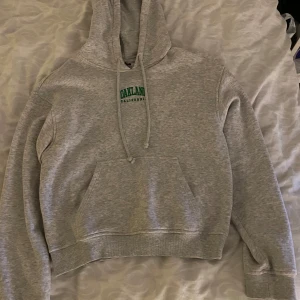 Grå hoodie med tryck - Säljer en grå hoodie med texten 'OAKLAND' i grönt på bröstet. Tröjan har en klassisk känguruficka och justerbar huva med dragsko. Perfekt för en avslappnad stil.