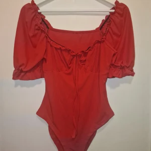 Röd body med puffärmar - Trendig röd body med puffiga ärmar och knytning framtill. Perfekt till middagen eller utekvällen när man vill sticka ut. Garderobsrensning så kika gärna på övriga auktioner.