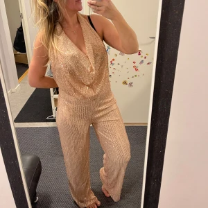Glittrig jumpsuit i guld - Snygg glittrig jumpsuit i guld med djup urringning och vida ben. Perfekt för en festlig kväll eller nyårsfirande. Den har en smickrande passform och ger en glamorös look.Helt ny