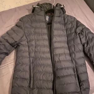 Svart Vinson Polo Jacka - Snygg svart dunjacka med huva och dragkedja. Perfekt för kyliga dagar med sin quiltade design och långa ärmar. Jackan har en stilren look och är ett måste för garderoben.