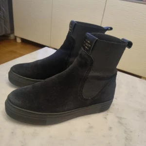 Svarta mocka boots - Snygga svarta boots i mocka med en stilren design. De har en högre skaft och en diskret logga på framsidan. Perfekta för en trendig look.
