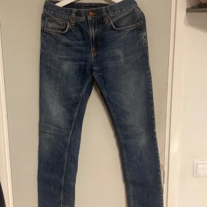 Blå jeans från Nudie Jeans - Snygga blå jeans från Nudie Jeans med klassisk femficksdesign och knappgylf. De har en stilren look med orange sömmar och en bekväm passform. Perfekta för en avslappnad stil. Jeansen har ett hål i höger framficka som enkelt går att laga. Byxorna är i modellen Gritty Jackson och i färgen Blue Slate. Storleken är W28 och L32.