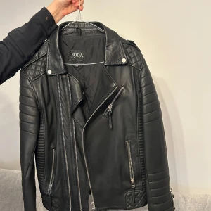 Svart skinnjacka från Boda Skins - Snygg svart skinnjacka från Boda Skins med coola detaljer som dragkedjor och quiltade axlar. Perfekt för en edgy look. Jackan har långa ärmar och en klassisk bikerstil. Storlek M