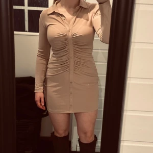 Beige klänning med knappar - Snygg beige klänning med långa ärmar och knappar framtill. (3 översta är riktiga resten fasta) Åtsittande passform med rynkdetaljer längs med knäppningen som ger en hour glass effekt. Helt ny från chiquelle, säljer pga att den aldrig kmr till användning. Storlek L men tror den passar M också. 