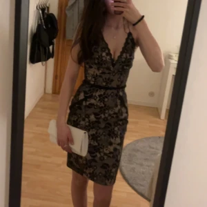 Svart spetsklänning från Kardashian Kollection - Elegant svart spetsklänning från Kardashian Kollection med djup v-ringning och figurnära passform. Klänningen har en ljusrosa grund med svart spets över. Öppen rygg med liten slits. 