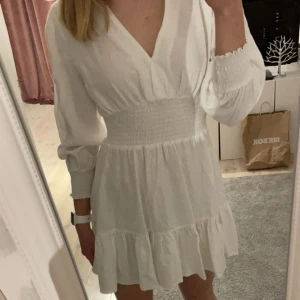 Vit kortklänning med v-ringning - Säljer en söt vit kortklänning med v-ringning och långa ärmar. Klänningen har en smockdetalj i midjan och volang nedtill. Perfekt sommar- eller studentklänning!