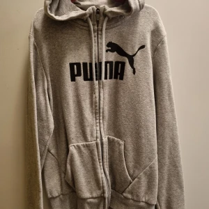 Puma - Säljer en grå hoodie från Puma med dragkedja och huva. Den har en stor Puma-logga på framsidan och praktiska fickor. 