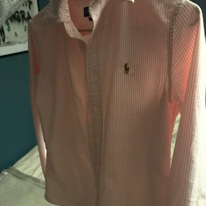  Ralph Lauren skjorta - Säljer en stilren rosa och vit randig skjorta från Ralph Lauren. Skjortan har en slim fit passform och är långärmad med knappar framtill. Perfekt för en klassisk och elegant look.