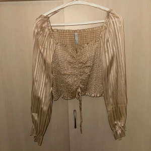 Guld randig blus - Snygg guldig blus med randigt mönster och långa ärmar. Blusen har en smockad design med knytning framtill och är perfekt för en stilren look.