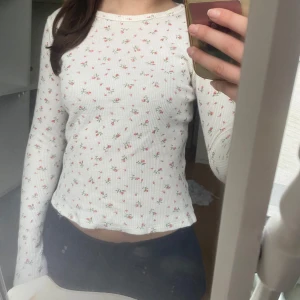 Vit blommig topp från Brandy Melville - Säljer en super söt vit åtsittande topp från Brandy Melville med små blommor i rosa och rött. Toppen är ribbad och har långa ärmar. Den är i fint skick! .