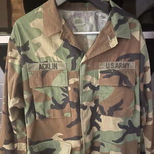 Camouflage militärjacka - Säljer en cool militärjacka i klassiskt camouflage-mönster. Jackan har två stora bröstfickor med lock och knappar, samt texten 'U.S. ARMY' och 'ACKLIN' på bröstet. Perfekt för en avslappnad stil.