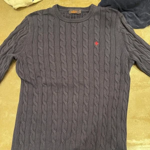 Mörkgrå stickad tröja från Morris - Säljer en stilren mörkgrå stickad tröja från Morris med kabelstickat mönster. Tröjan har en liten röd broderad detalj på bröstet och rund halsringning. Perfekt för kyliga dagar!